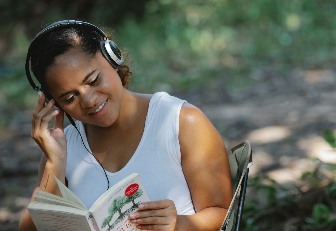 Photo of a smiling woman wearing headphones and reading a book outside